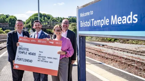 Councilor Mike Bell, MP for North Somerset Sadik Al-Hassan, West of England Mayor Helen Godwin and leader of Bristol City Council Tony Dyer at Bristol Temple Meads station. Helen and Sadik are smiling and holding a large railway ticket, with stations listed.