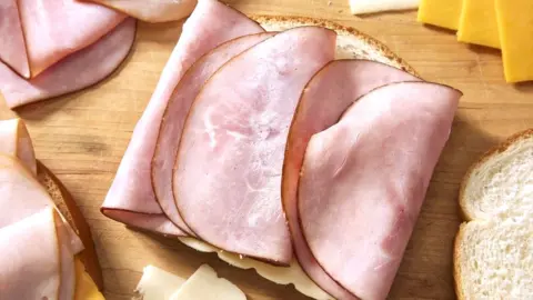 Getty Images Three slices of ham folded on white bread with slices of cheese and bread surrounding it