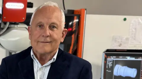Delta Fire Ian Gardner wearing a suit jacket and a white shirt stood in front of a computer screen and what appears to be the arm of a machine