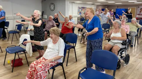 Men and women are taking part in an exercise class in a hall. Some are standing and some are seated and one woman is in a wheelchair. They are following the instructor's arm movement. The hall is nearly full and there is a clock on the wall, with a noticeboard.