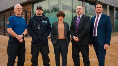 Essex Police Five people stand in a line - a man wearing a royal blue top and black trousers, a police officer in all black with a black cap, a woman in a dark suit with a light brown top, a man in a black suit with a striped tie and a man in a blue suit with a purple tie.