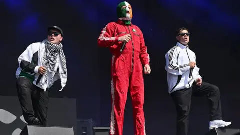 A man in a red boiler suit and tricolour balaclava stands between two men who wear white tracksuit tops. The man on the left holds a black microphone and wears a black and white scarf and a black baseball cap. The man on the right has on black sunglasses and white trainers