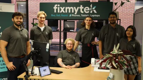 FixMyTek Six people, all in FixMyTek polo shirts, in the office. There is a sign in the background which says "Fixmytek - buy, sell, repair - phones, tablets, laptops" and the group are gathered around a desk which has a computer, a tablet and a large plant on it. 