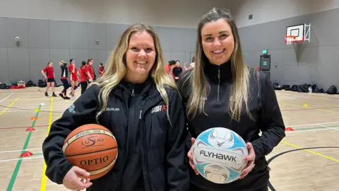 BBC Libby and Laura stand indoors on a sports court, each holding a ball. Libby, on the left, holds a basketball, and Laura, on the right, holds a netball. Both wear dark sports jackets. Behind them, several groups of people in red sports uniforms practice on the court, which has marked lines, cones, and a basketball hoop on the wall.