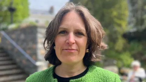 A woman with short brown hair, with gold earring and a green cardigan looks at the camera. 