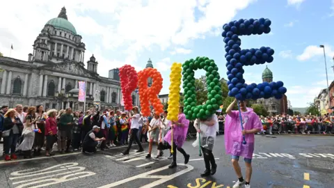 PA Media People taking part in the Belfast Pride parade in the city centre in 2023 on the street, people in pink t-shirts are holding balloons that read PRIDE in red, orange, yellow, green and blue. They are marching outside city hall with crowds watching on. 