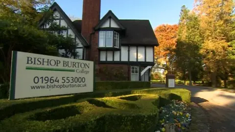 A Tudor style building surrounded by neatly trimmed hedges, colourful flowers and large trees. A sign in the foreground reads Bishop Burton College.