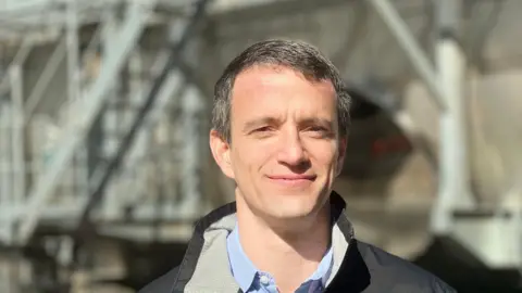 Close up of Nelson, who is smiling and has grey hair. He is wearing a blue shirt and a black coat and standing outside in front of a blurred background.