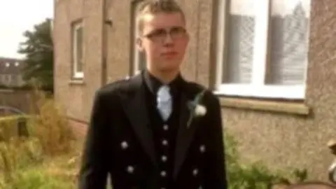 Police Scotland A young man with short hair and glasses wearing a jacket and tie, stands outside