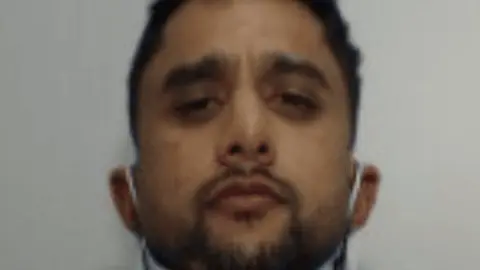A mugshot of a youngish man with dark hair and a dark beard. He had a face mask around his ears that is pulled down to reveal his face. 