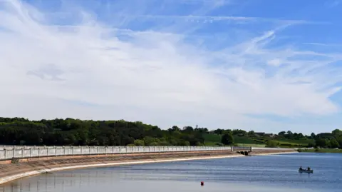 A wide view of Blithfield Reservoir in the middle of the countryside. A causeway is running along the left of the reservoir, with trees in the background. Two people can be seen in a boat on the water on the right.