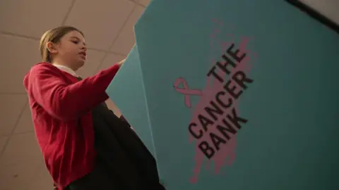 A young schoolgirl, wearing a red cardigan, and grey dress, puts something into a blue box which has 'The Cancer Bank' written on the side of it, in pink and black lettering 