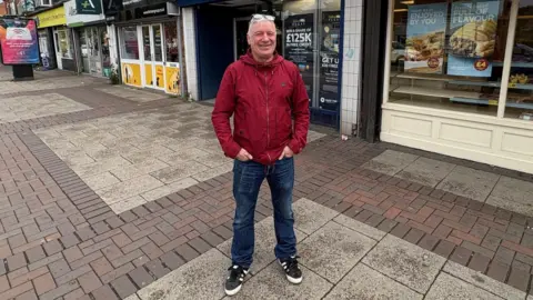 A man in his 50s smiles for the camera. He is wearing a red jacket and navy jeans and black trainers. Behind him is a parade of shops.