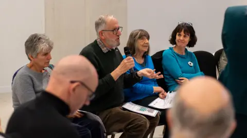 Emma Doyle/University of Sheffield Three women and one man are sat on chairs facing to the right. The man is wearing glasses and speaking into a microphone while gesturing with his other hand. He has some papers on his knee. Two men are sat facing him in the foreground.