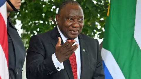 South Africa's President Cyril Ramaphosa, in red tie and grey suit, gestures with his hand and smiles outside the White House between the US and South African flags - 21 May 2025.