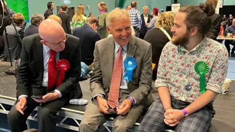 Charlotte Rose/BBC Three male candidates (left to right) with red Labour, blue Conservative and Green rosettes sit on a stage in a busy counting hall.