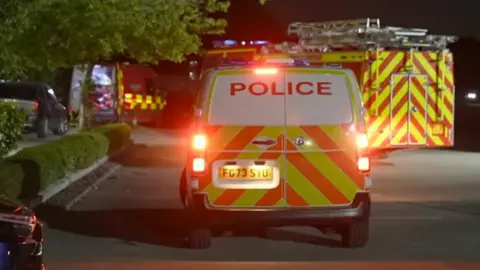 The rear of a police van in a car park with yellow and orange chevrons on the back and its lights flashing with a fire engine behind it