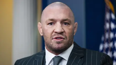 EPA-EFE/REX/Shutterstock A close-up shot of Conor McGregor as he stands in the White House press briefing room. The background is blurred. He is wearing a green and white pinstripe suit, a white collared shirt and a green patterned tie. 