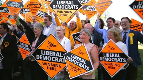 Councillors outside gathered together holding signs saying "Liberal Democrats winning here".
