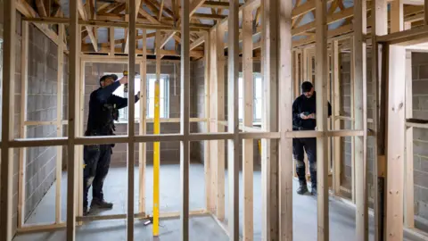 A stock picture of two men working in a house that is partly built with wooden beams put up to divide future rooms.