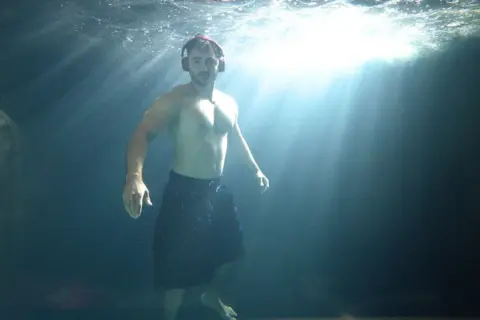 Simon Lodge A man in a kilt with headphones on underwater with light bursting through from above