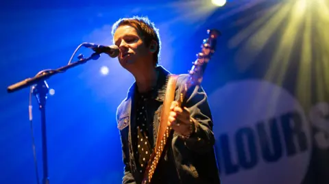 Raymond Meade, on stage with an Ocean Colour Scene backdrop, sings into a microphone while playing bass guitar. He is wearing a navy and white spotted shirt and a denim jacket.