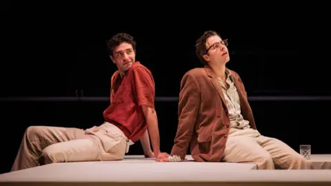 Mark Senior Two men sit on a stage. The man on the left has dark hair and wears an orange shirt and light-coloured trousers. The man on the right has dark blond hair and wears a brown suede jacket and a light-coloured shirt and trousers.