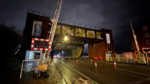BBC Drypool Bridge in Hull. The structure has two raised barriers to show it is open and a number of orange cones on a tarmac road. The bridge is silhouetted against the night sky and street lights. The metal and brick structure is painted yellow and white.