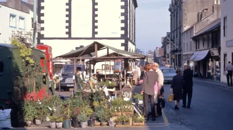 Keswick Museum People gather around a market stall selling plants in Keswick. There are shops behind it, as well as a couple of cars. The quality of the picture is slightly faded.