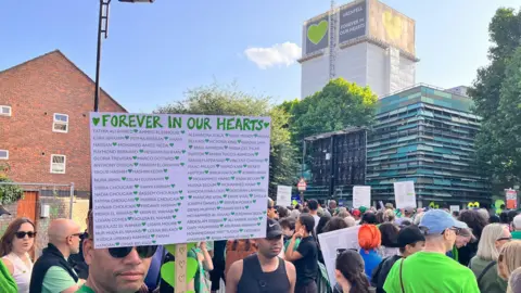 Silent walk on the eighth anniversary of the fire, crowds wearing green and holding placards, with Grenfell Tower in the background.