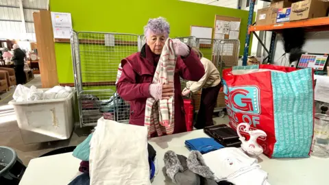 Martin Giles/BBC A woman wearing a burgundy coat is sorting through bags of clothes. There are cages against a green wall behind her.