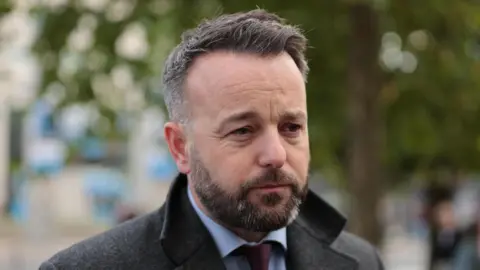 PA Media Foyle SDLP MLA Colum Eastwood stands outside a courthouse. He is wearing a blue shirt, red tie and heavy winter coat. He has short greying black hair.