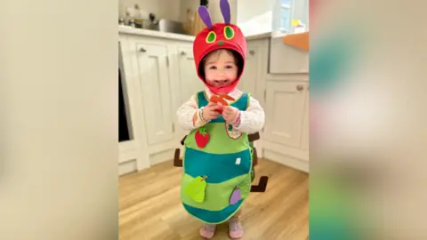 Maya is dressed in a Very Hungry Caterpillar costume and is smiling for the camera and is standing in what appears to be a kitchen, with an oven behind her.