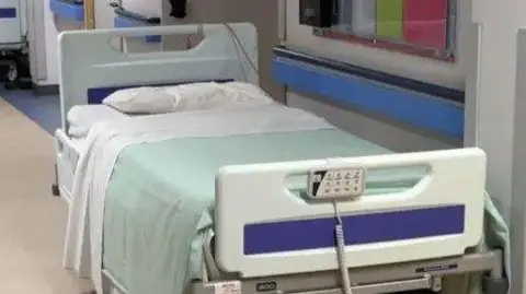 A hospital bed, which has white pillows and sheet and a pale green blanket on it.