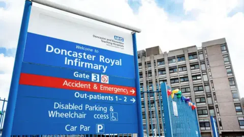 Doncaster and Bassetlaw Teaching Hospitals NHS Foundations Trust A large blue sign with white writing. Behind the sign is a large tower block with several floors and windows