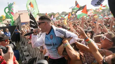 Getty Images Moglai Bap of Kneecap, a young man with a skin-fade haircut and wearing sunglasses, yells into his microphone as he performs with his back to the crowd, who are close enough to grab his arms.