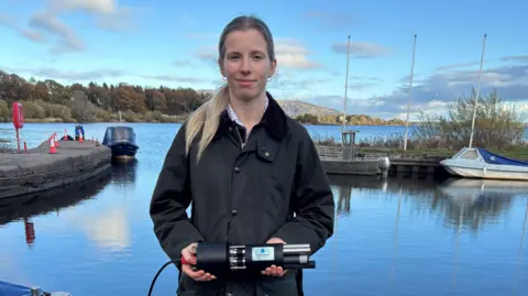University of Exeter Kathryn Thomas has blonde hair tied back in a ponytail. She is wearing a green coat and holding a black testing device. She is standing in front of a Scottish lock and looking at the camera, and smiling.