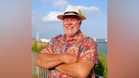 Tony Francis Tony Francis stands with his back to some railings and the sea. He is wearing a Panama hat and a colourful short-sleeved shirt. He is smiling broadly and has a grey/white beard.