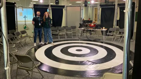 BBC Prime's communications director, Jaime Pontins and Grow Up's co-director, Erin Fletcher. stand behind the shows round black and white stage which resembles an archery target. It is surrounded by fold-out chairs with black curtains behind them