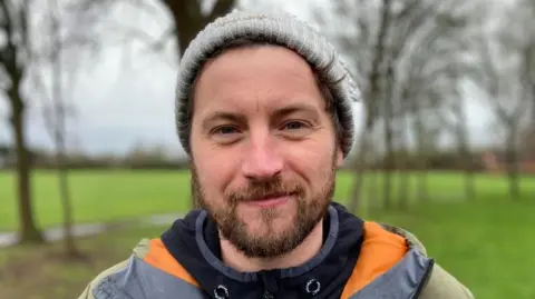Helen Mulroy/BBC Rob Ford stands in a park and looks directly at the camera. He has brown hair and a beard and wears a grey beanie and an orange, grey and black coat. 