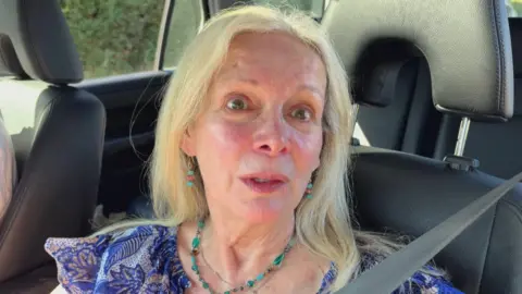 Woman with green necklace and blue top in front seat of cat with seatbelt on