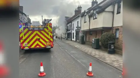 Cambridgeshire Fire and Rescue The picture is looking at the right side of the street. Parked up on the left is a fire engine, that is red and yellow. One of the buildings on the right has grey smoke coming out of it. 