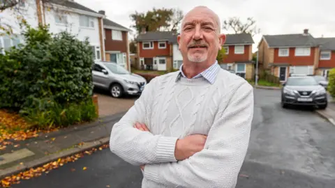 People's Postcode Lottery A mature man wearing a white jumper smiling to camera standing in a cul de sac with cars and houses behind him