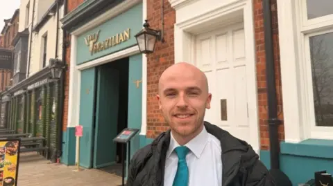 The Brazilian is painted in gold letters on a turqouise background. Markus wears a matching turquoise tie, a white shirt and black coat and smiles for the camera.