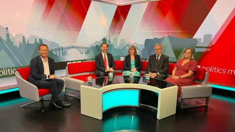 A man sits in a red chair in a TV studio, while two men and two women sit next to him on a red sofa. The screen behind them says Politics Midlands.