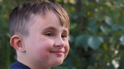 A close-up image of a young boy's face. He is looking away from the camera but smiling. There are green leaves from a tree in the background. 
