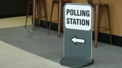 A sign saying 'polling station' with an arrow pointing to the left. Behind it, three chairs are visible.