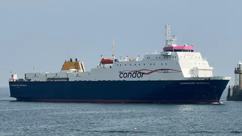 BBC A large commercial vessel, the Commodore Goodwill, heading into a harbour. Skies are blue.
