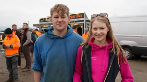 Shaun Whitmore/BBC A young man and woman stand side by side smiling at the camera. The man on the left is wearing a blue hoodie and has blonde hair which hangs down onto his forehead. The woman has long hair with sunglasses on her head. She is wearing a pint hoodie with a gilet jacket over the top. 