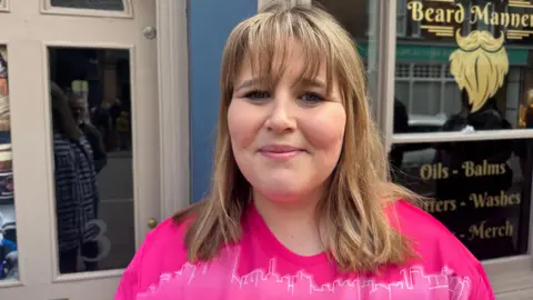 Aimee Dexter/BBC Woman with blond hair wears a pink T-shirt and stands with her back to a pub